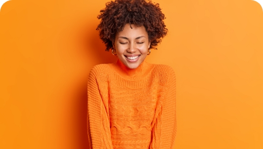 cheerful-beautiful-woman-closes-eyes-smiles-broadly-wears-long-sleeved-sweater-poses-against-vivid-orange-wall-has-happy-smile-face